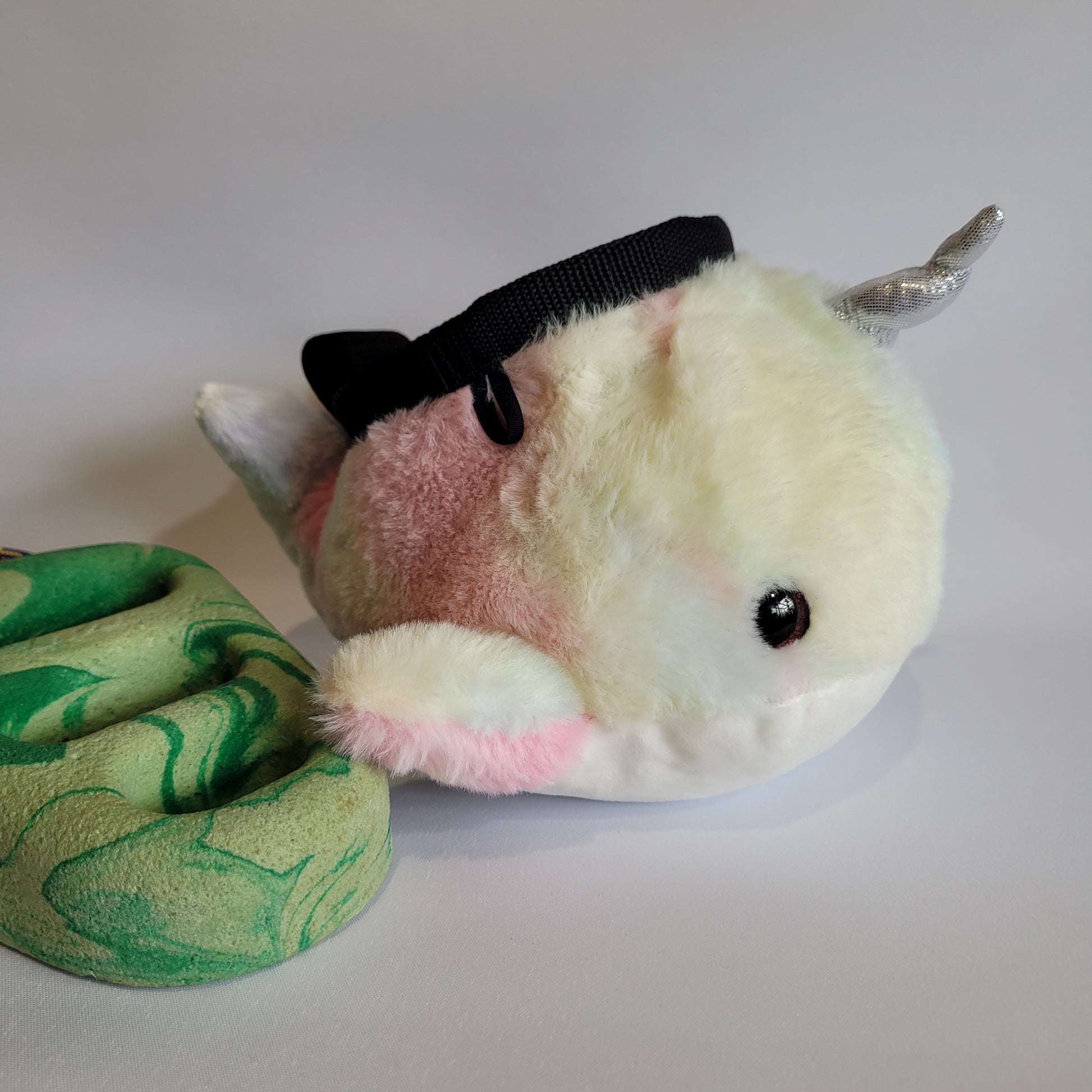 A plush rock climbing chalk bag designed to look like a pastel-colored narwhal, featuring a shiny silver horn, small fins, and a soft gradient of pink, white, and cream fur. The bag has a black drawstring closure on top and is positioned next to a green marbled climbing hold.