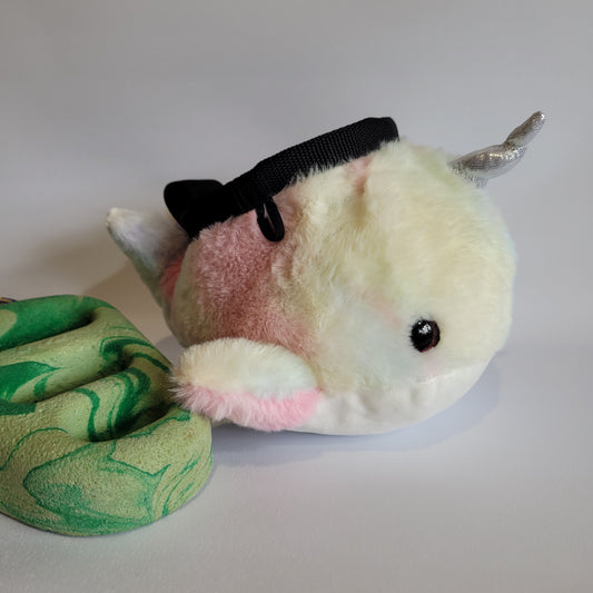 A plush rock climbing chalk bag designed to look like a pastel-colored narwhal, featuring a shiny silver horn, small fins, and a soft gradient of pink, white, and cream fur. The bag has a black drawstring closure on top and is positioned next to a green marbled climbing hold.