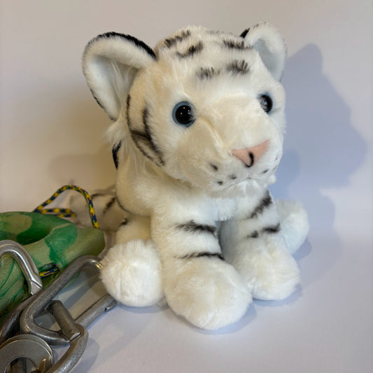 Plush white tiger chalk bag with black stripes, blue eyes, and a pink nose, sitting upright next to green climbing gear and carabiners on a white background.